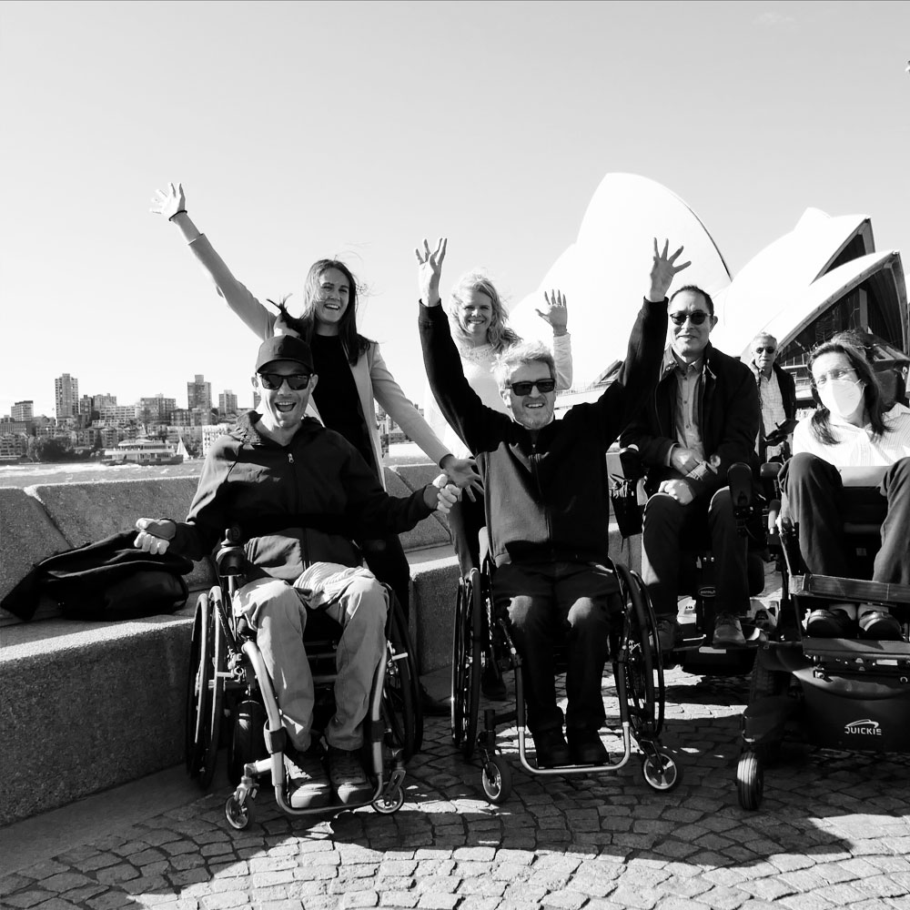 A range of people with disabilities smiling with hands in the air, Sydney Opera House in the background