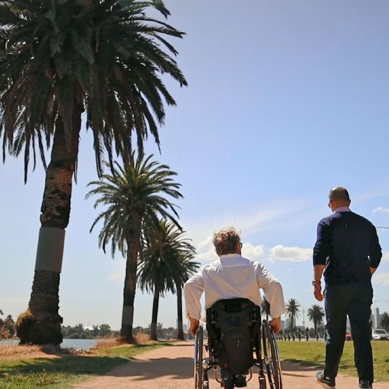 the silhouettes of 2 people facing away from the camera, one in a wheelchair on walking. The sky is bright blue, they're next to a lake