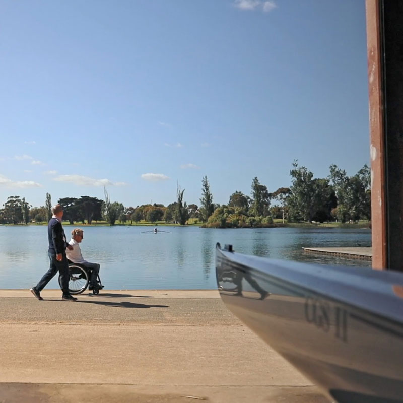 a photo of 2 people walking next to a lake on a sunny day. One's in a wheelchair wearing a white shirt and jeans, the other has dark blue slacks and a blue jumper